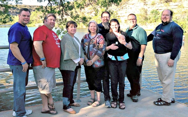 Eight members of the Gardner Seminar on a lakeside dock, one holding a baby, a limestone bluff behind them